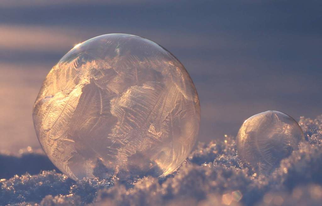 a soap bubble resting on a snowy surface, with the surface of the bubble partially covered in icy tentacles - the sun is shining from behind and creating a lovely visual effect